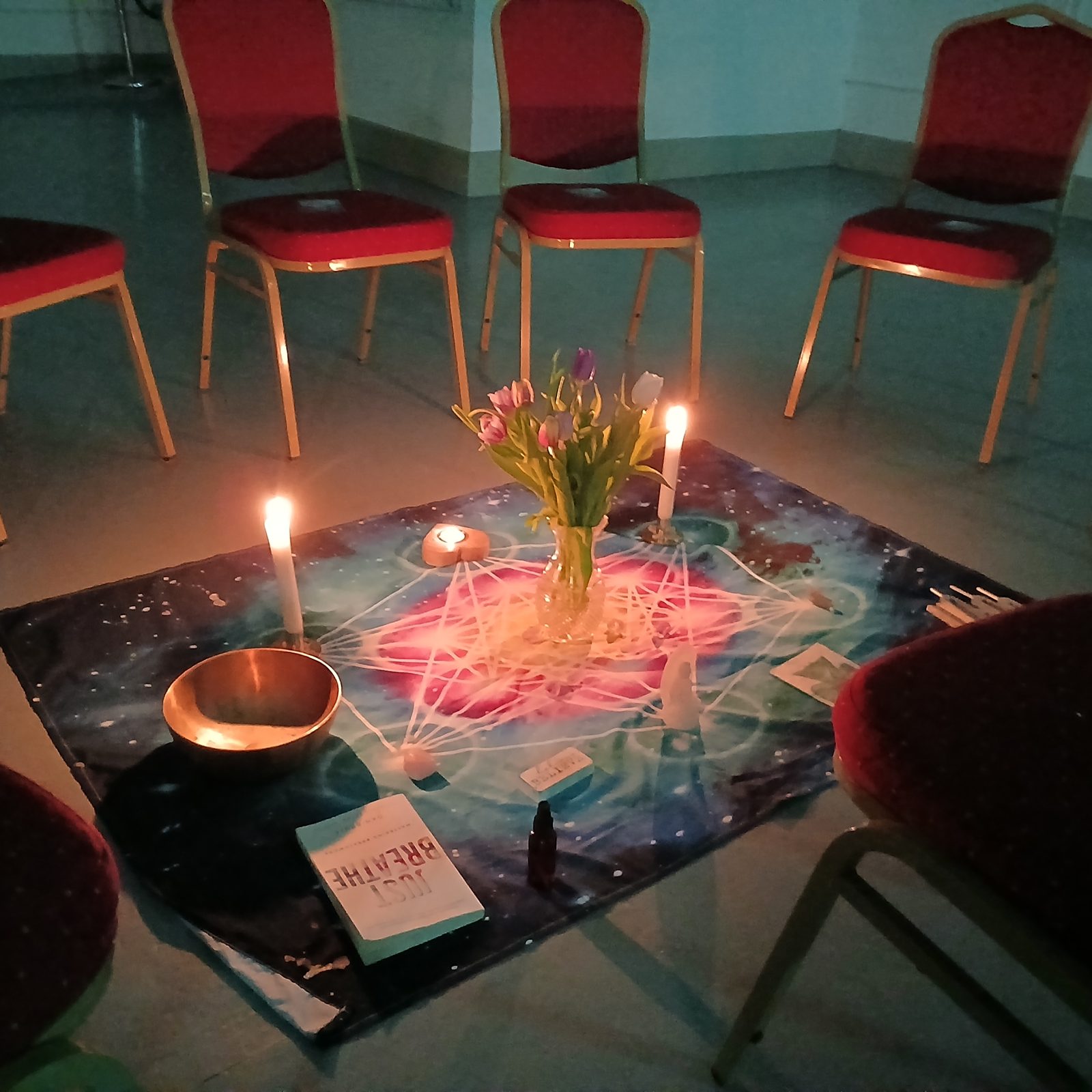 Sound bath ceremony setup — chairs arranged in a circle around a candlelit altar cloth with tulips, singing bowl and crystal grid, ready for participants